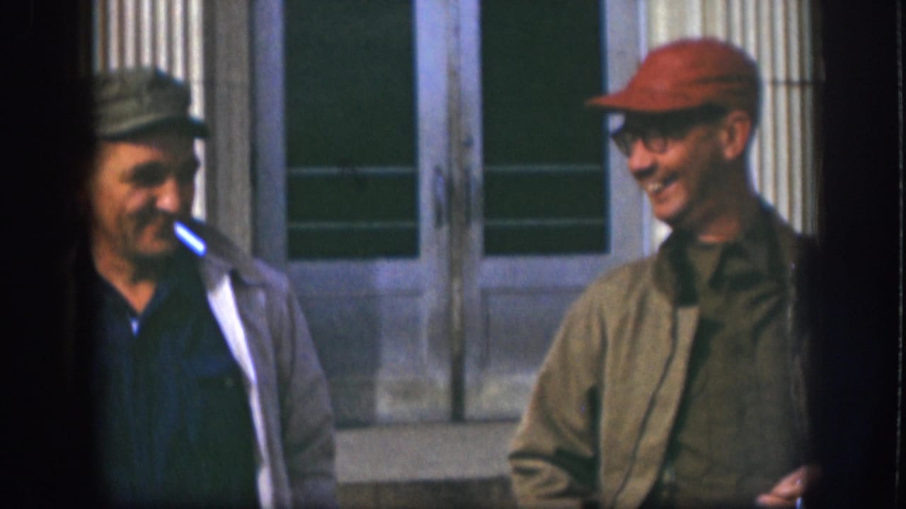 In 1958 Kansas home-movie footage, two adult men in caps stand near a columned building while one smokes a cigarette.