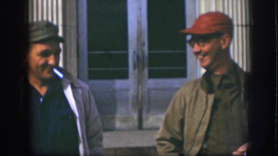 In 1958 Kansas home-movie footage, two adult men in caps stand near a columned building while one smokes a cigarette.
