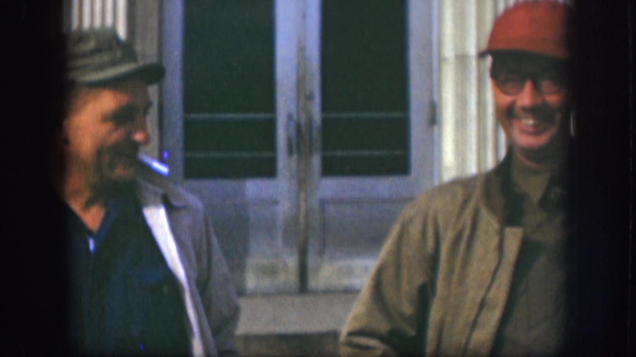 Two adult men in caps, one wearing glasses, appear to smile and talk outside a building in 1958 Kansas footage.