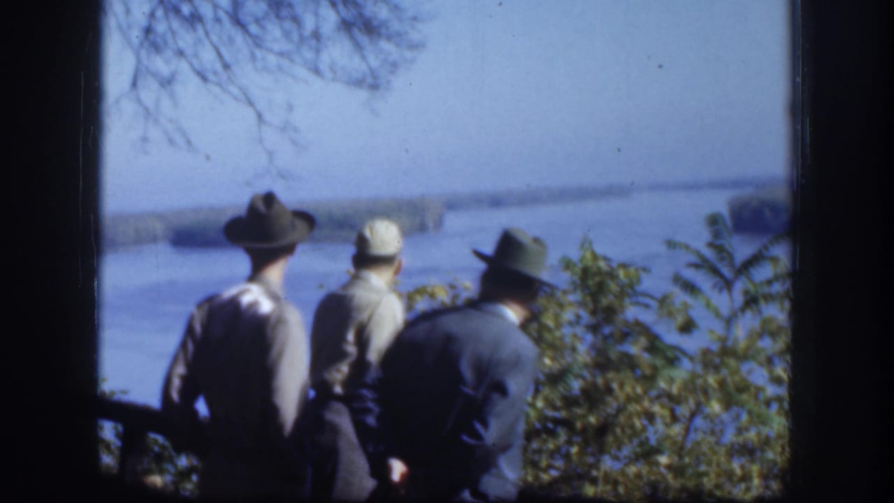 Three adult men in hats stand near trees and look over a broad river or lake in 1958 Kansas home-movie footage.