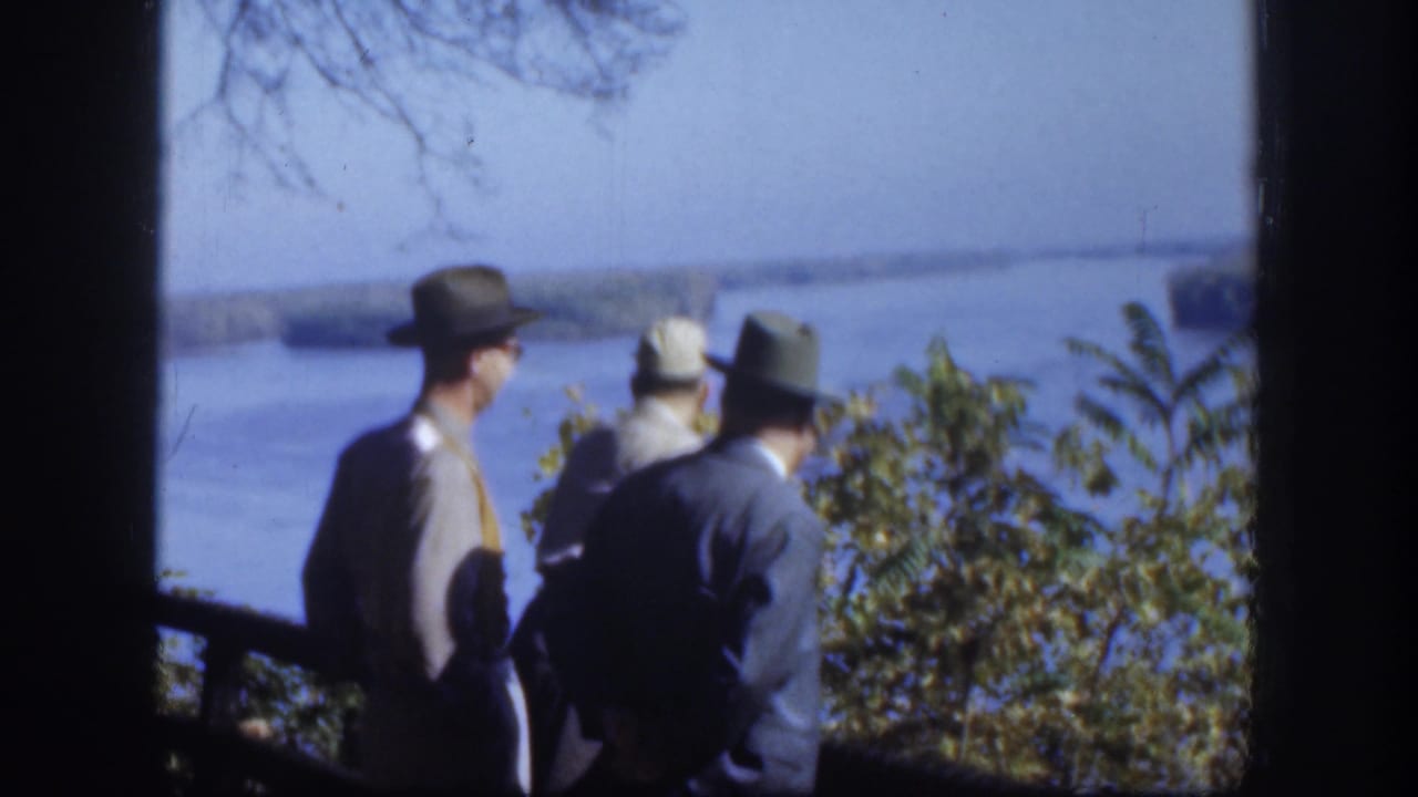 In 1958 Kansas home-movie footage, three men in brimmed hats face a broad river from a shaded overlook.