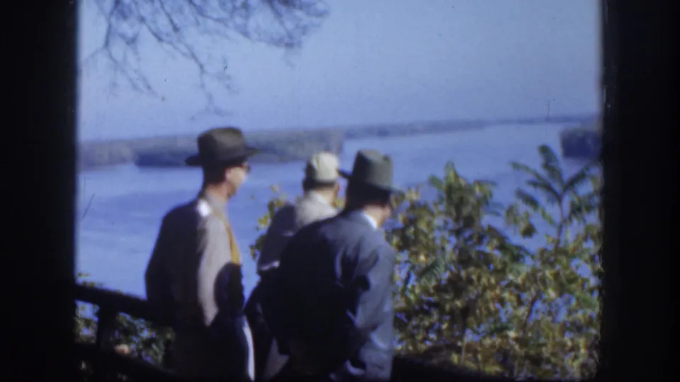 In 1958 Kansas home-movie footage, three men in brimmed hats face a broad river from a shaded overlook.