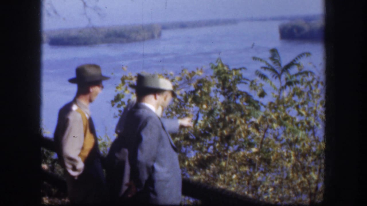 Two hat-wearing men are visible at a Kansas river overlook, with one gesturing toward the water in the distance.