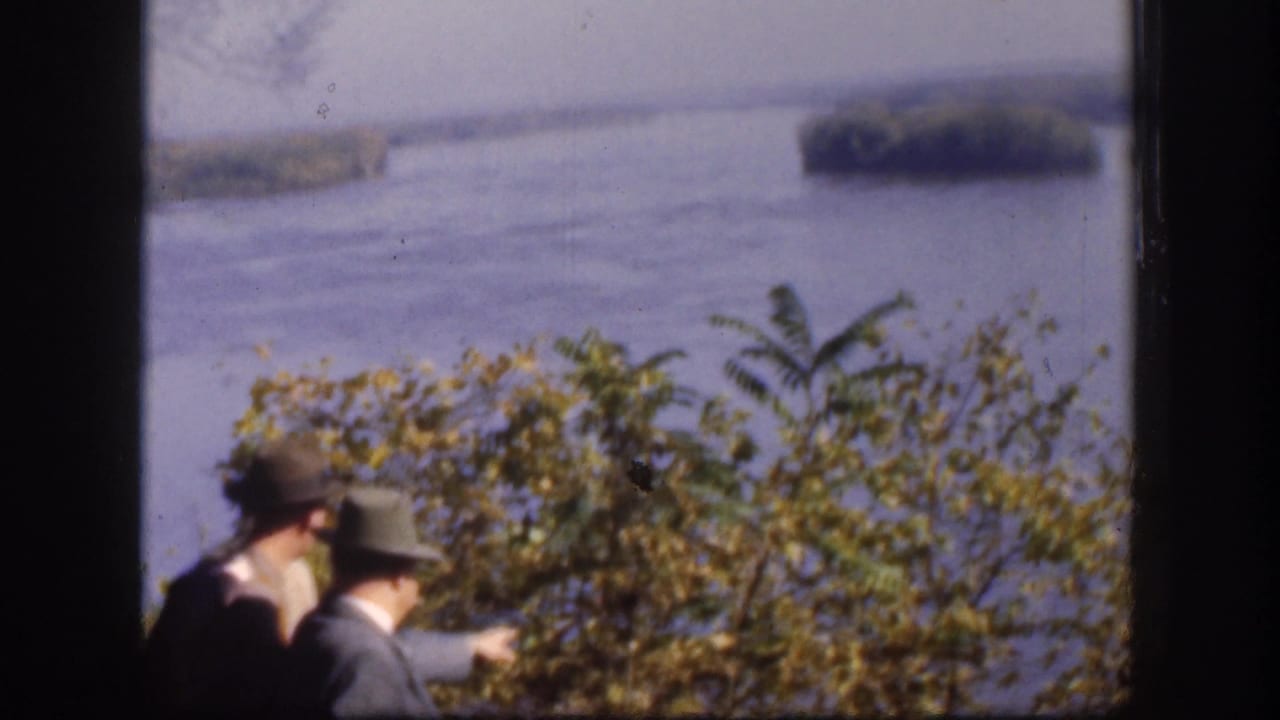 Partly cropped figures in hats remain at the lower edge as the camera favors shrubs and the river beyond.