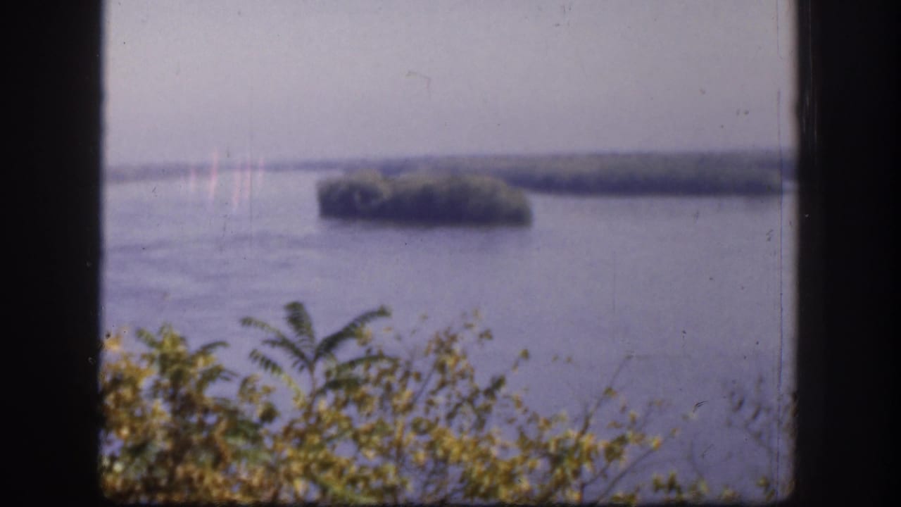 1958 Kansas 8mm frame showing a broad river or lake with small wooded islands beyond foreground vegetation.