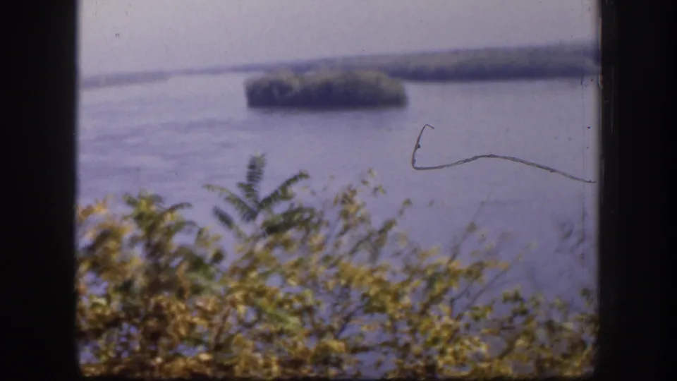 1958 Kansas 8mm frame of a hazy river or lake vista with shrubs below, a wooded island beyond, and visible film wear.