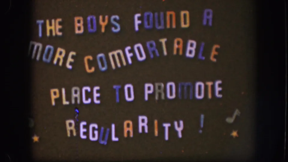 1958 Kansas home-movie frame with the handmade sign centered, showing playful multicolored lettering and small decorative symbols.