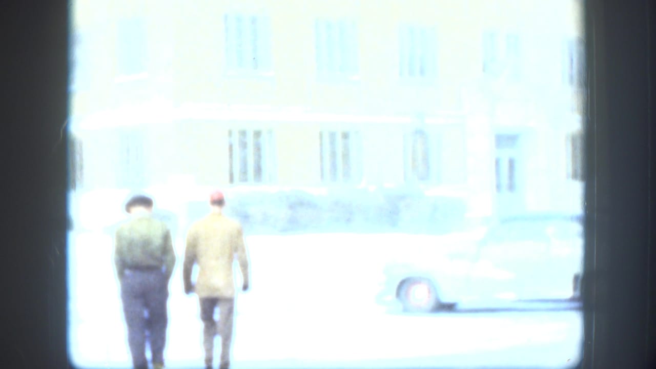 In Kansas home-movie footage from 1958, two men walk away from the camera near a pale building and parked car on a snowy day.