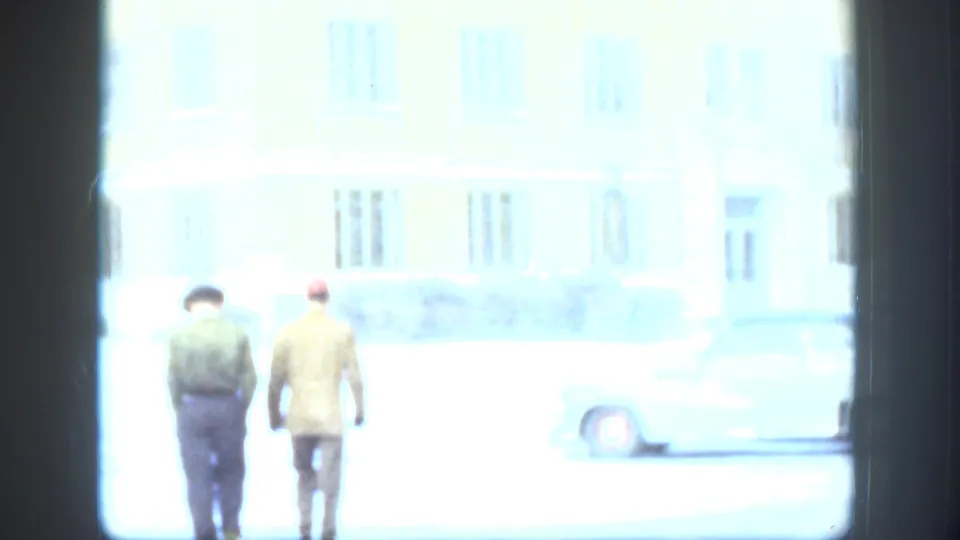 In Kansas home-movie footage from 1958, two men walk away from the camera near a pale building and parked car on a snowy day.