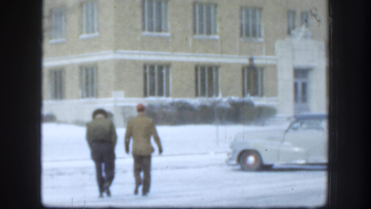 Two men in winter clothing walk away across a snowy foreground toward a yellow brick building in Kansas, with a light-colored car nearby.