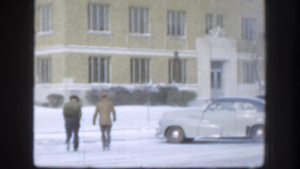 The 1958 Kansas scene shows two men walking in snow before a yellow institutional-looking building and a rounded light-colored car.