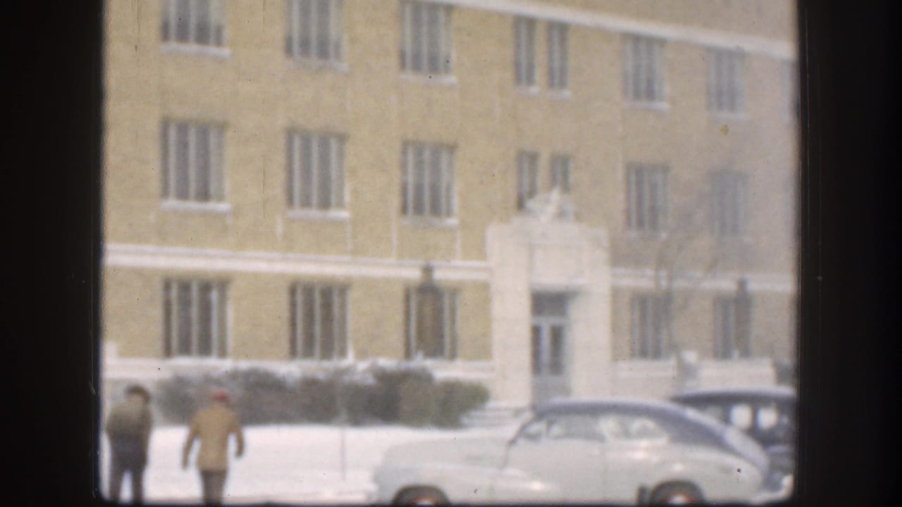 Near the end of the clip, two men appear low in frame beside a light-colored car, with the yellow Kansas building behind them.