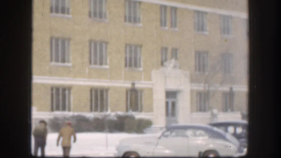 Near the end of the clip, two men appear low in frame beside a light-colored car, with the yellow Kansas building behind them.