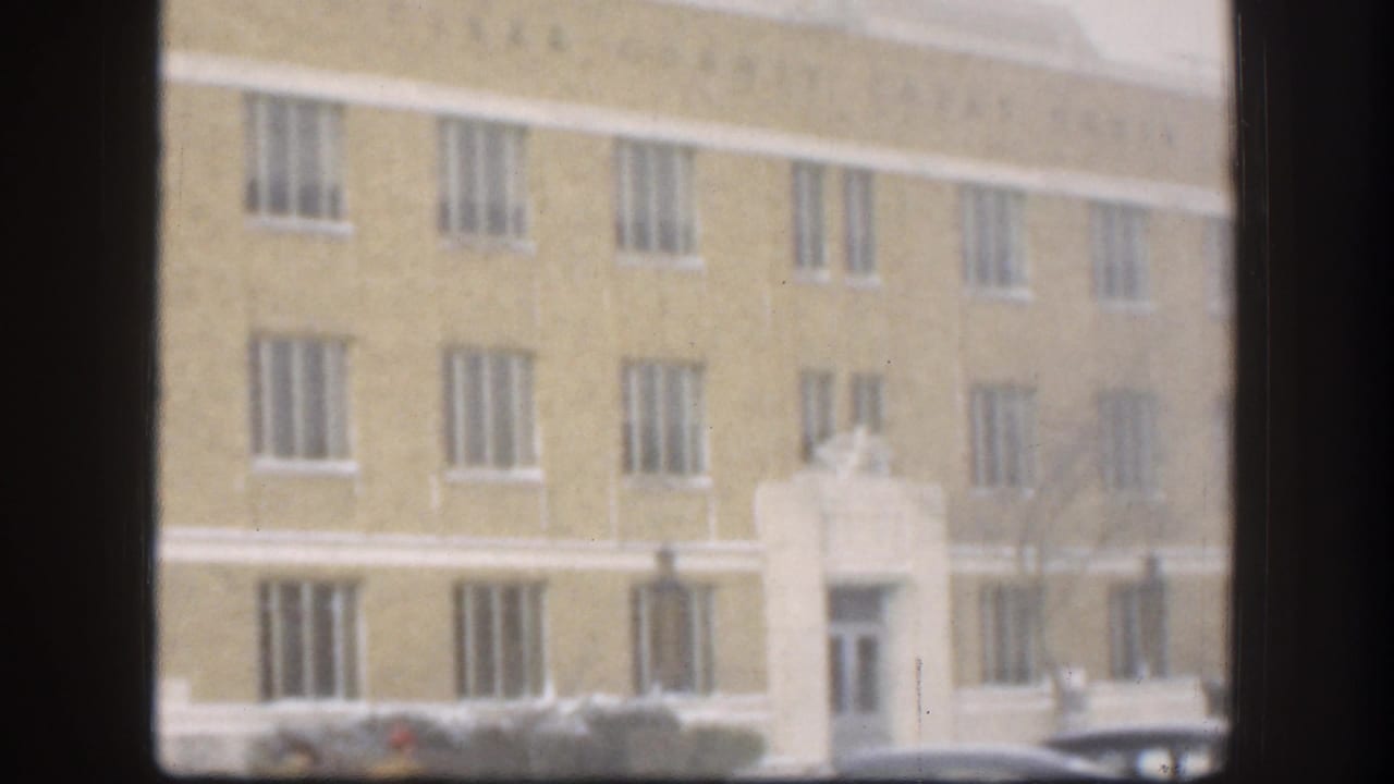 1958 Kansas 8mm frame showing a pale yellow institutional facade with barred windows and a white central entrance, seen through dark foreground muntins.