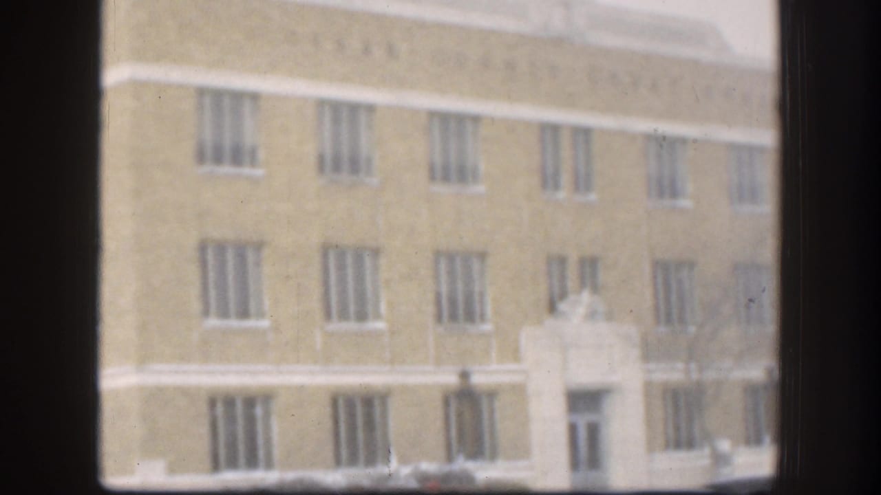 1958 Kansas home-movie frame with rows of narrow barred windows on a yellowish institutional facade, partially blocked by dark window framing.