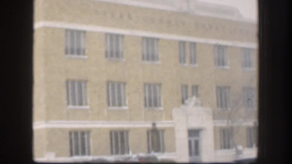 1958 Kansas home-movie frame with rows of narrow barred windows on a yellowish institutional facade, partially blocked by dark window framing.