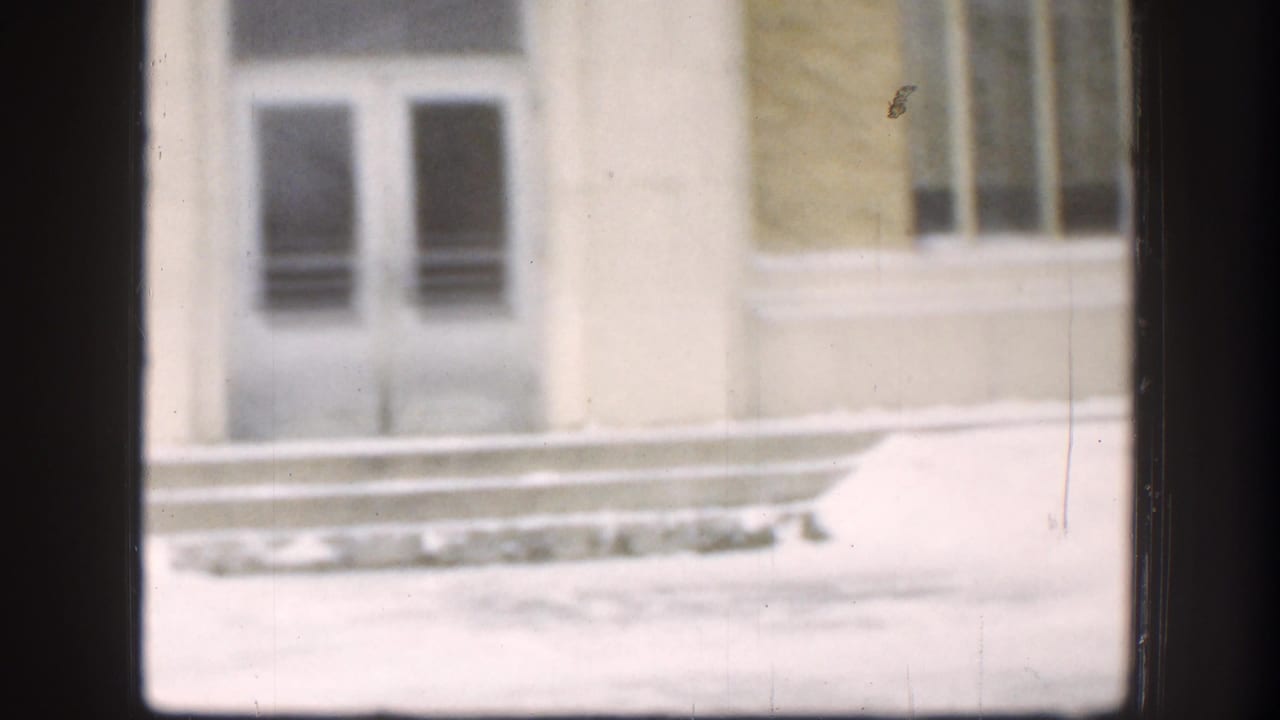 Soft 1958 Kansas 8mm view of the building entrance area, with white masonry, dark doors, barred windows, and snow piled along the base.