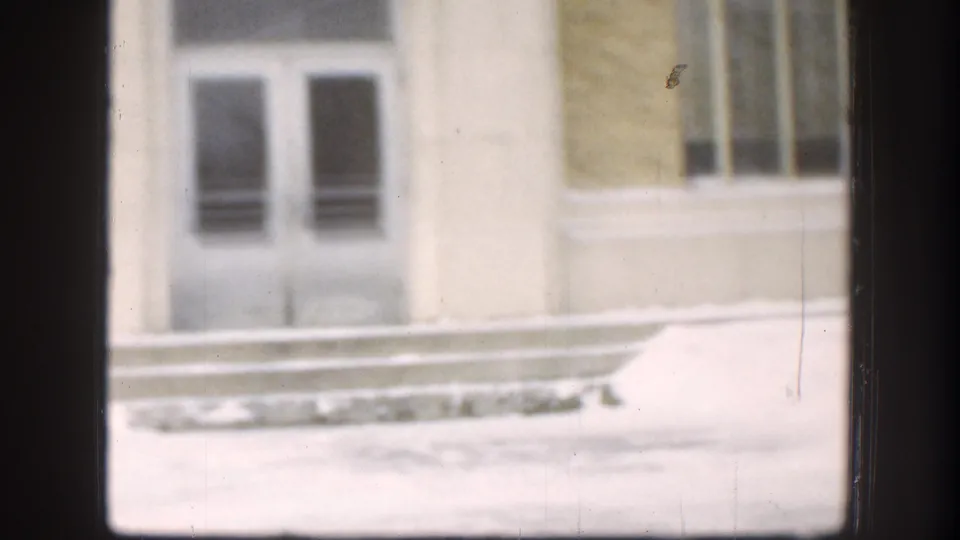Soft 1958 Kansas 8mm view of the building entrance area, with white masonry, dark doors, barred windows, and snow piled along the base.