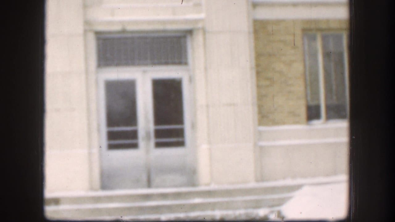 1958 Kansas frame showing the white-framed entrance, barred transom area, yellowish facade, and adjacent windows, viewed through a dark interior window frame.