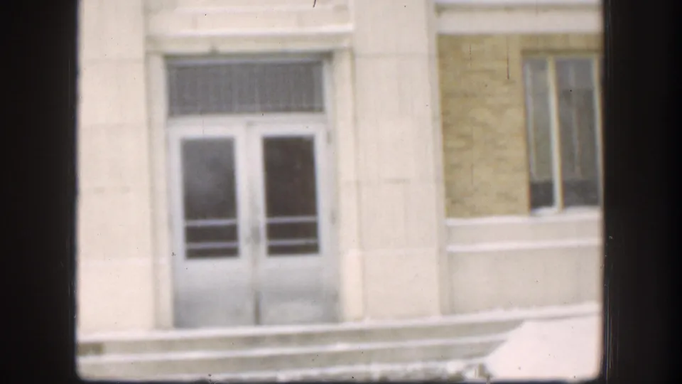 1958 Kansas frame showing the white-framed entrance, barred transom area, yellowish facade, and adjacent windows, viewed through a dark interior window frame.