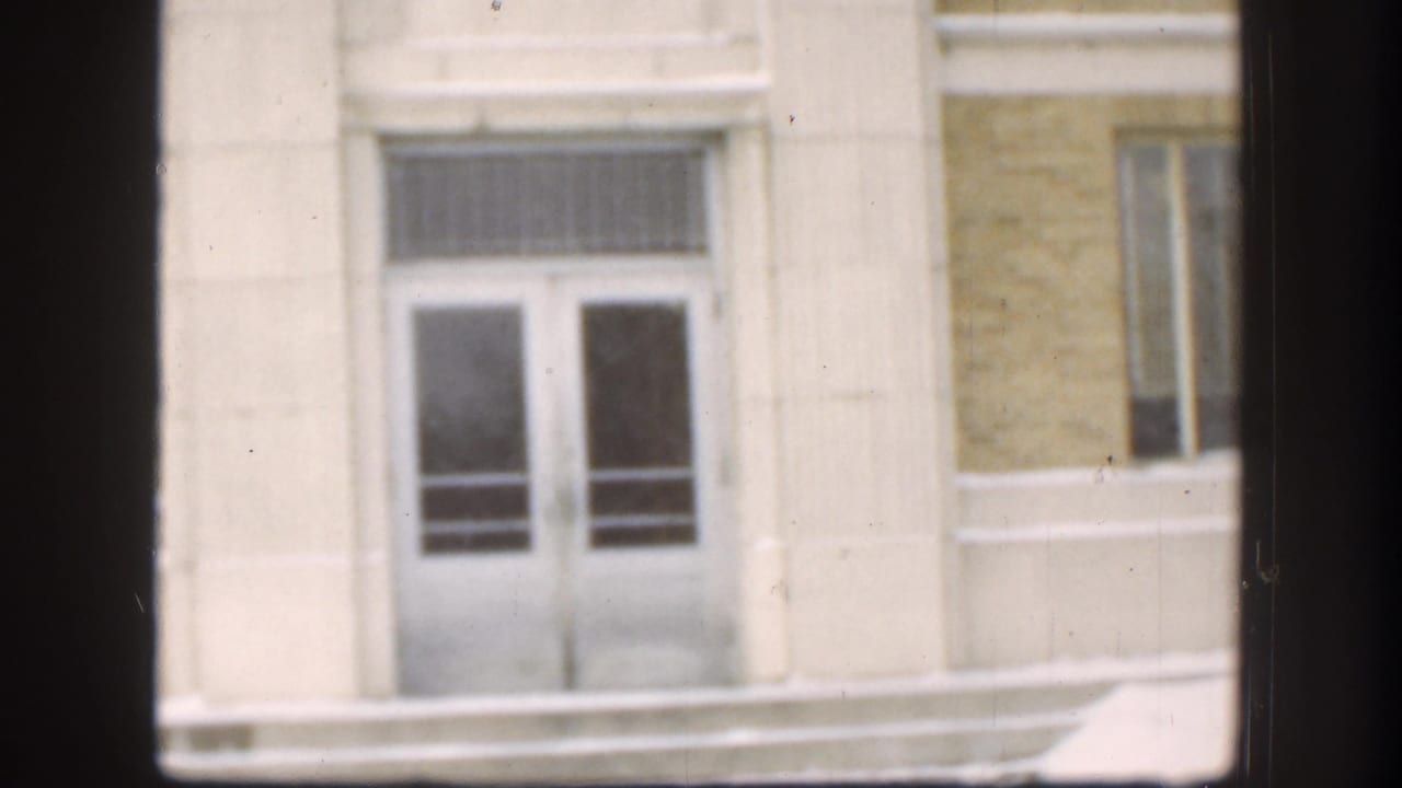 1958 Kansas 8mm frame showing a pale building facade, double doors, and snowy steps viewed through dark window mullions.