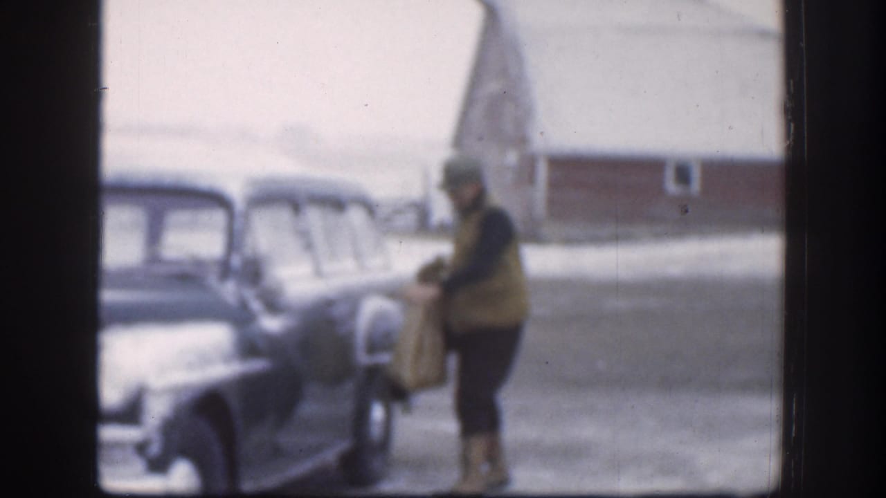 A man in a hat stands near a parked vintage car in a snowy rural setting.
