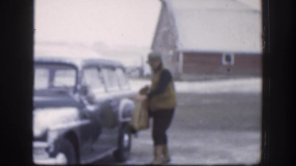 A man in winter clothing approaches a mid-century car in rural Kansas, with snow on the ground and a red outbuilding behind him.
