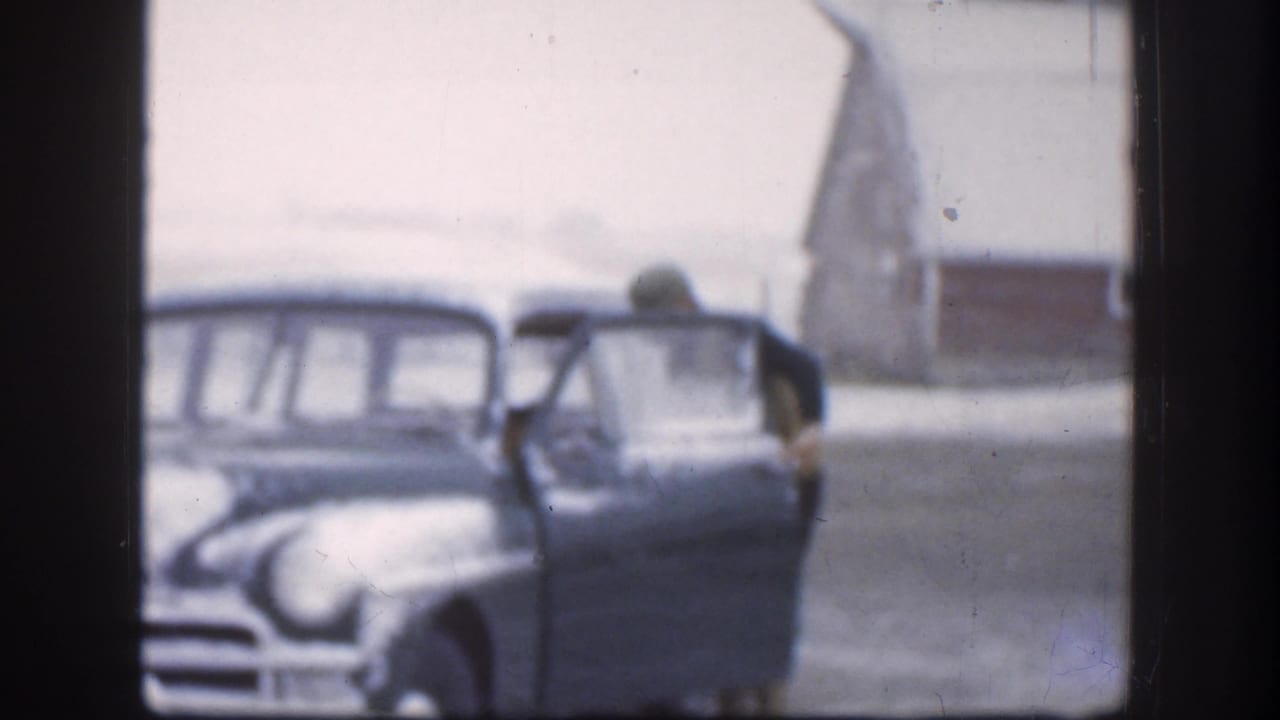 The man opens the sedan door in the Kansas winter scene, with the rounded car body and red outbuilding visible through soft 8mm focus.