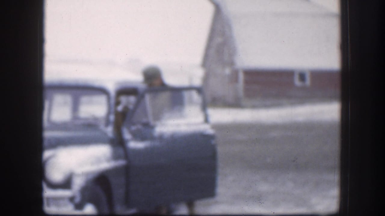 The man is partly hidden by the open sedan door in the 1958 Kansas home-movie frame, with a red outbuilding and snow visible behind him.