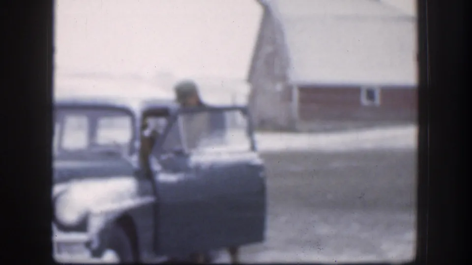 The man is partly hidden by the open sedan door in the 1958 Kansas home-movie frame, with a red outbuilding and snow visible behind him.