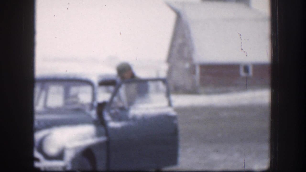 In 1958 Kansas home-movie footage, a man in winter clothing stands beside an open vintage car door with snow and a barn-like building behind him.