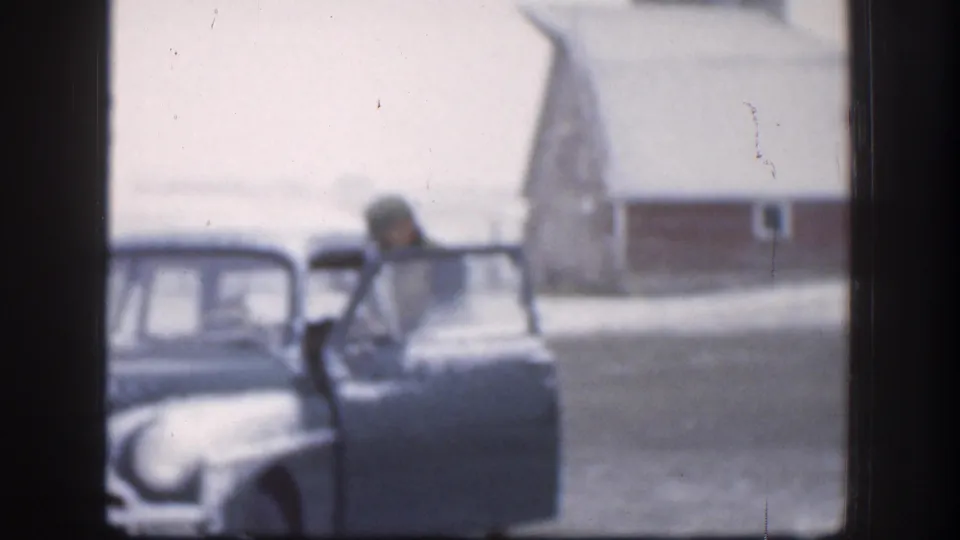 In 1958 Kansas home-movie footage, a man in winter clothing stands beside an open vintage car door with snow and a barn-like building behind him.