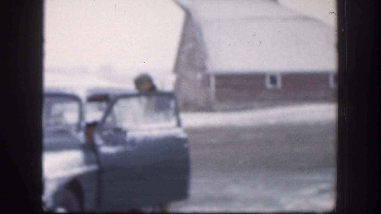 The man is partly visible near the open door of a vintage car in a snowy Kansas yard, with a red barn or outbuilding beyond the driveway.