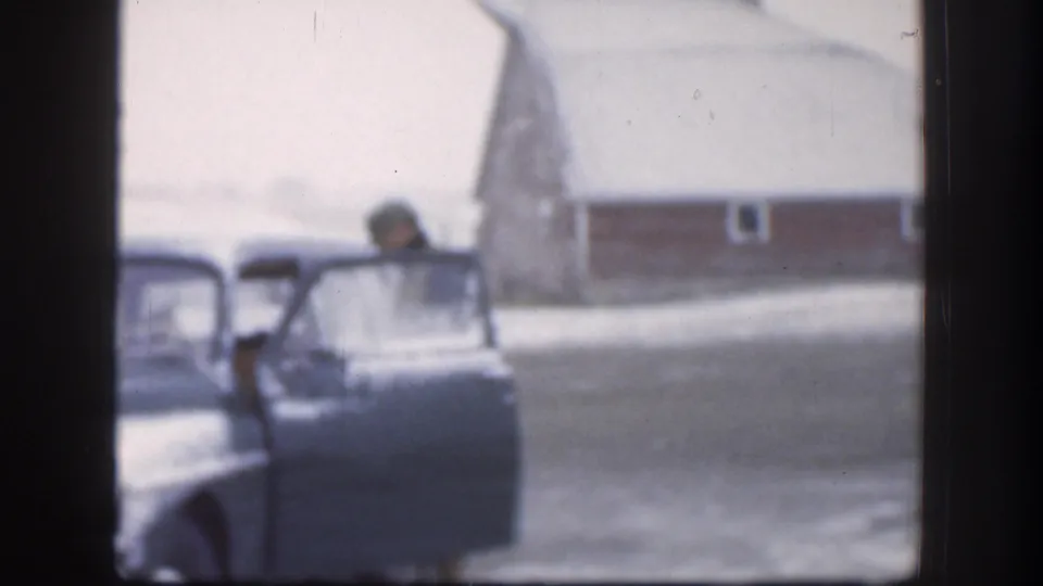 The man is partly visible near the open door of a vintage car in a snowy Kansas yard, with a red barn or outbuilding beyond the driveway.
