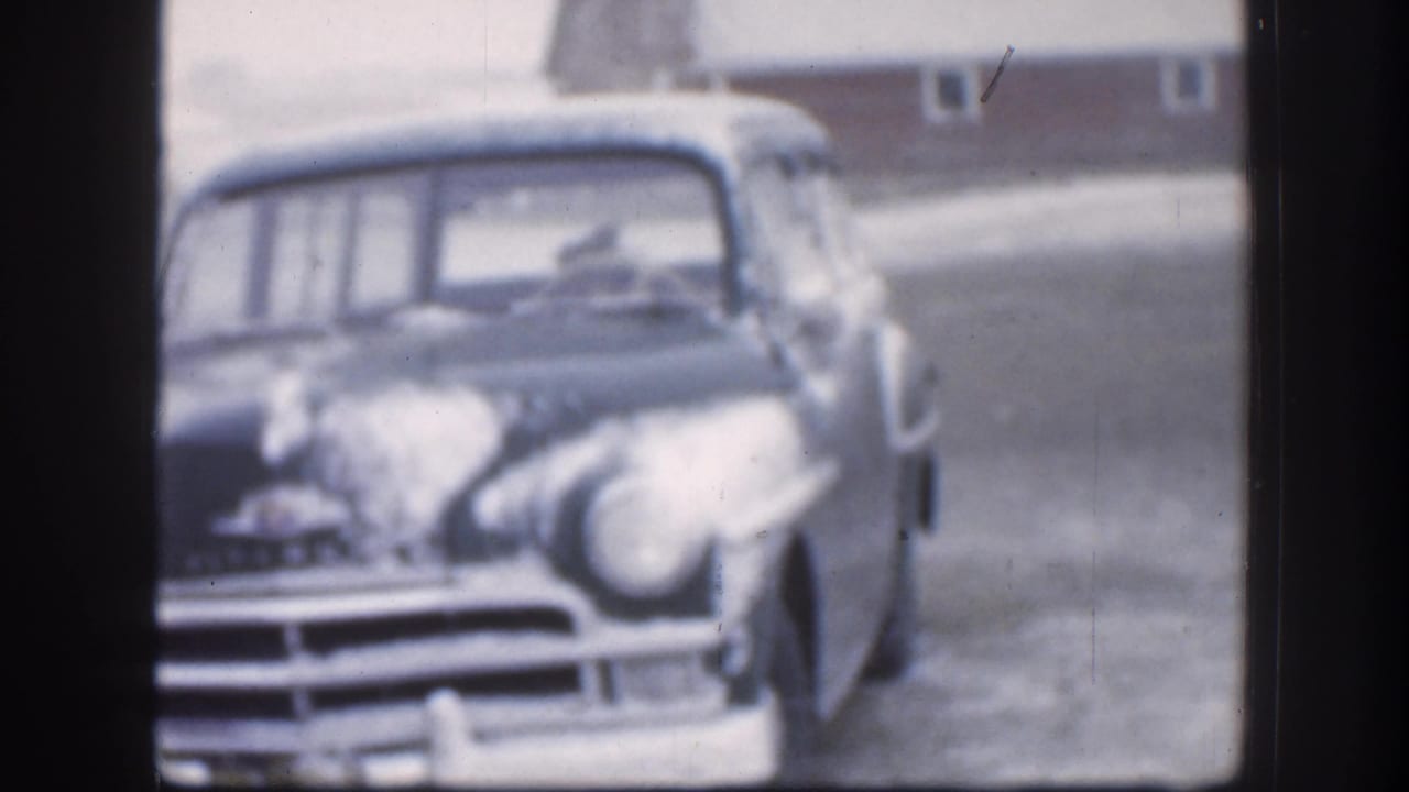 A snow-dusted vintage car faces the camera in the Kansas winter scene, with the driver area and barn-like building softly visible behind it.