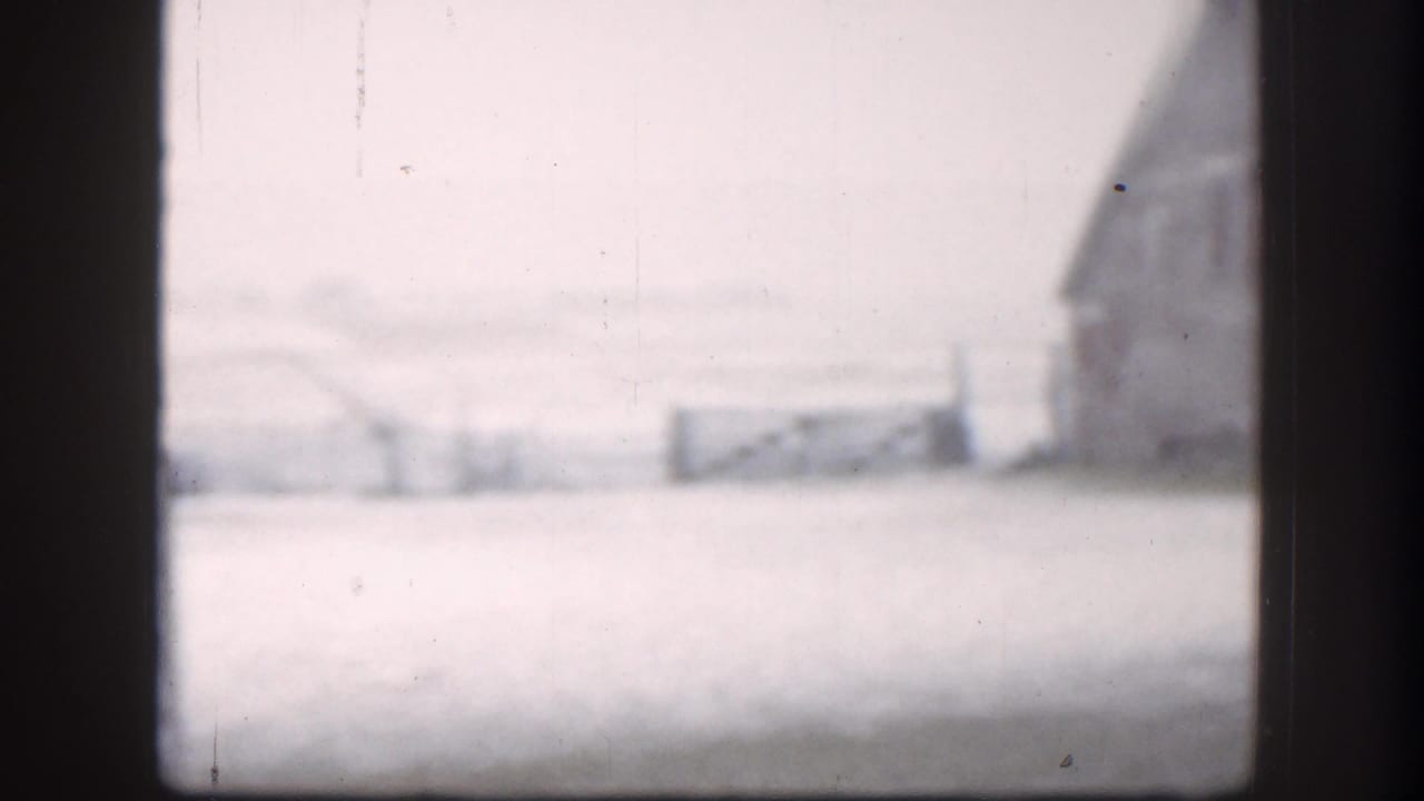 Snow-covered Kansas farmland with a distant barn-like building and pale sky, framed by dark vertical edges.