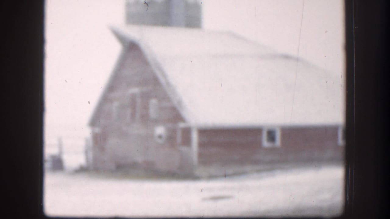 A large barn-like farm building stands beside a snowy Kansas yard or lane in soft 8mm footage.