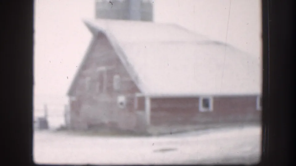 A large barn-like farm building stands beside a snowy Kansas yard or lane in soft 8mm footage.