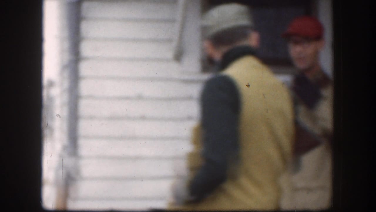 Two visible people near a white-sided farm building, including a man in a cap and vest, in 1958 Kansas home-movie footage.
