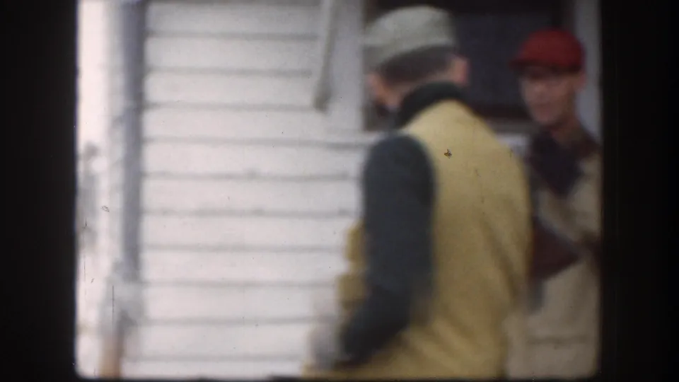 Two visible people near a white-sided farm building, including a man in a cap and vest, in 1958 Kansas home-movie footage.