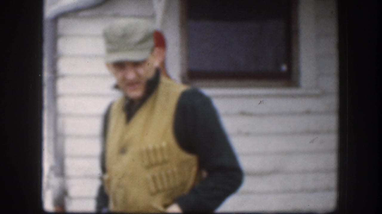 Adult man in a cap and beige hunting vest passes beside a white-sided house.