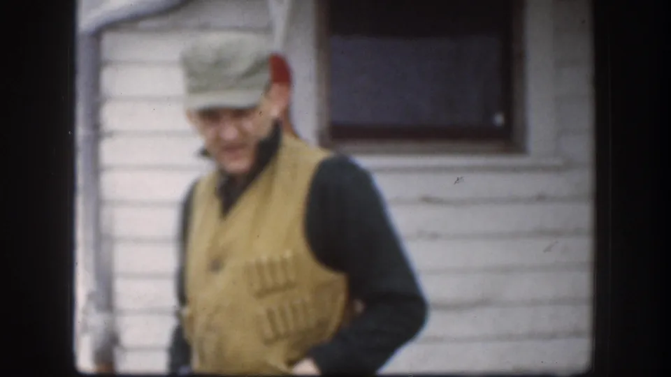 In this 1958 Kansas 8mm frame, an adult man in a cap and beige hunting vest moves past the camera beside a white-sided house.