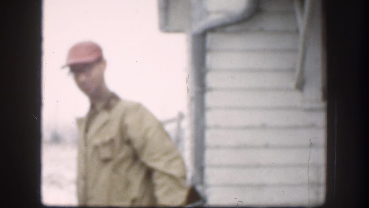 In the Kansas home-movie frame, an adult man in a red cap and tan jacket appears near the corner of a white-sided house.