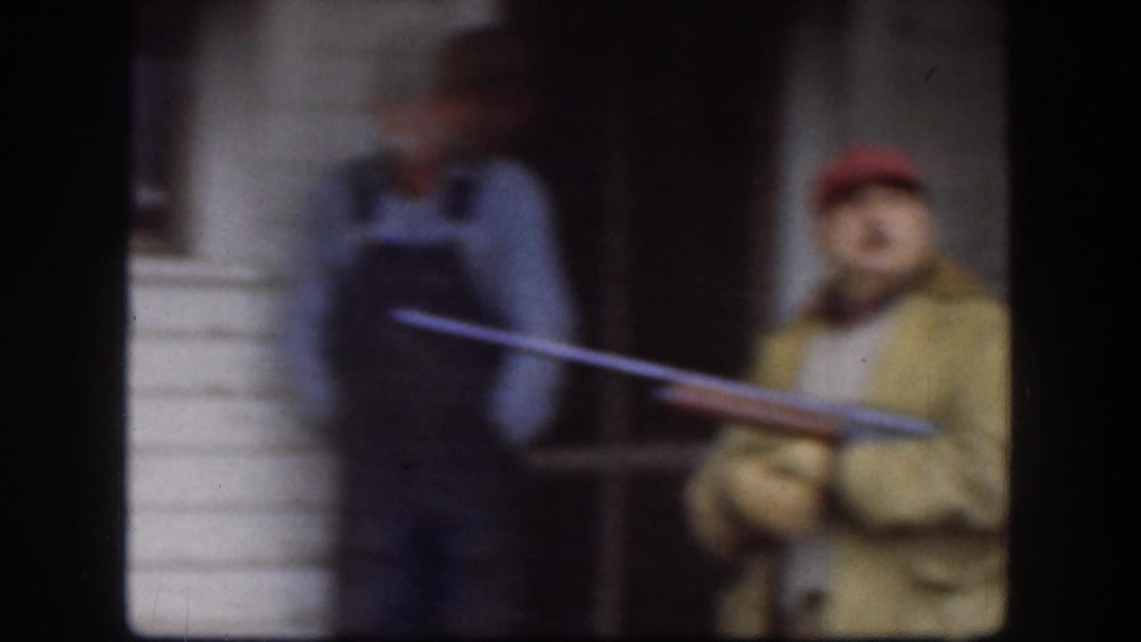 Man in red cap and yellow jacket holds a long gun-like prop on a porch near another person.