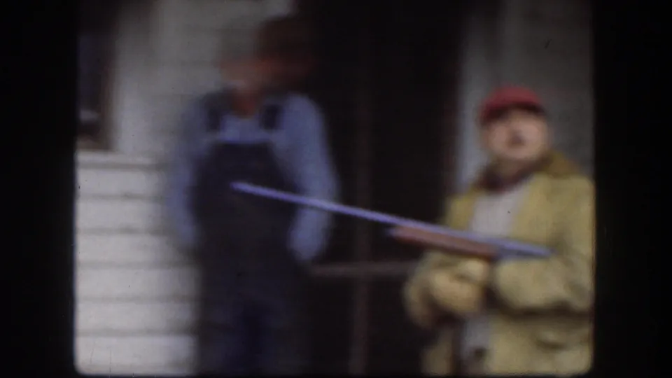 Kansas 1958 home-movie frame showing a red-capped man on a clapboard-house porch holding a long toy or replica firearm near a companion.