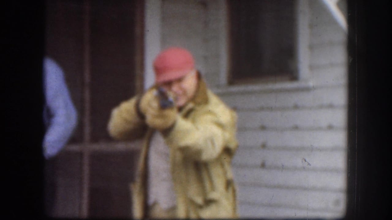 Kansas 1958 frame of a man in a red cap and yellow jacket aiming a toy or replica firearm toward the camera area on a porch.