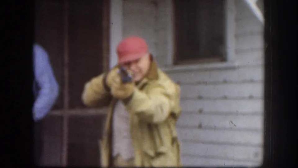 Kansas 1958 frame of a man in a red cap and yellow jacket aiming a toy or replica firearm toward the camera area on a porch.
