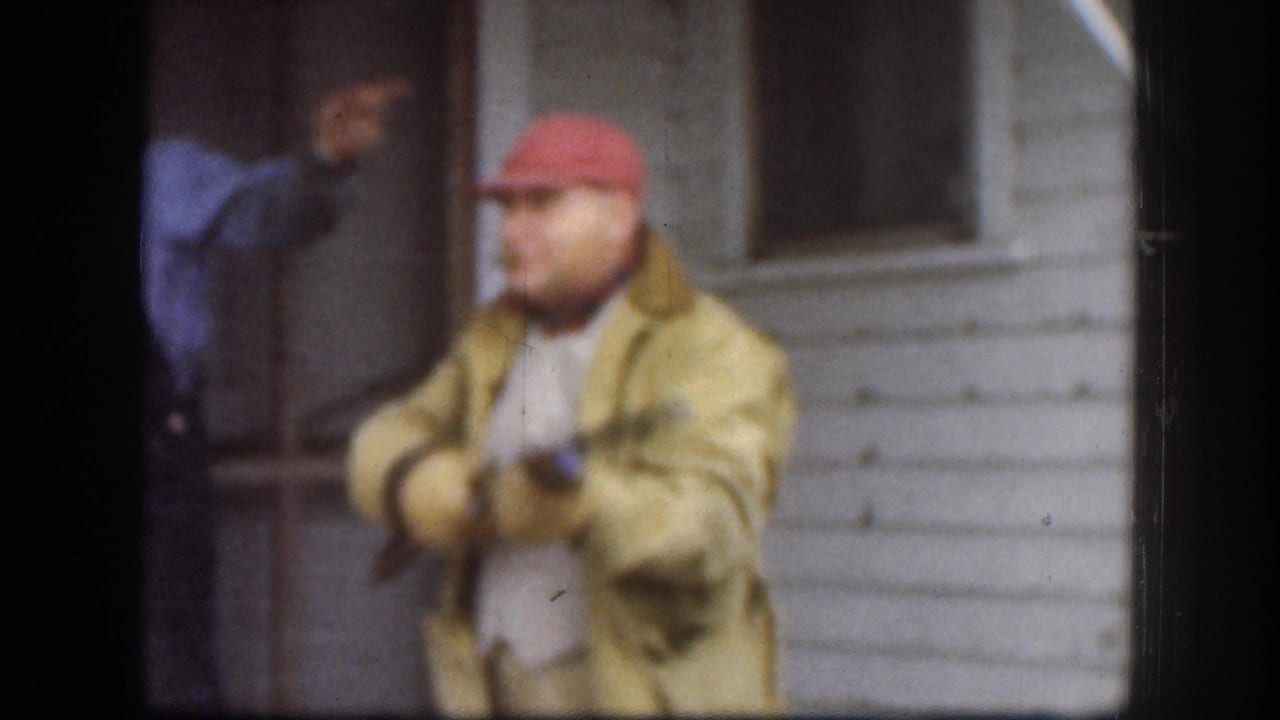 Soft 8mm Kansas 1958 frame showing the red-capped man holding a toy or replica firearm near a porch, with a companion partly visible behind him.