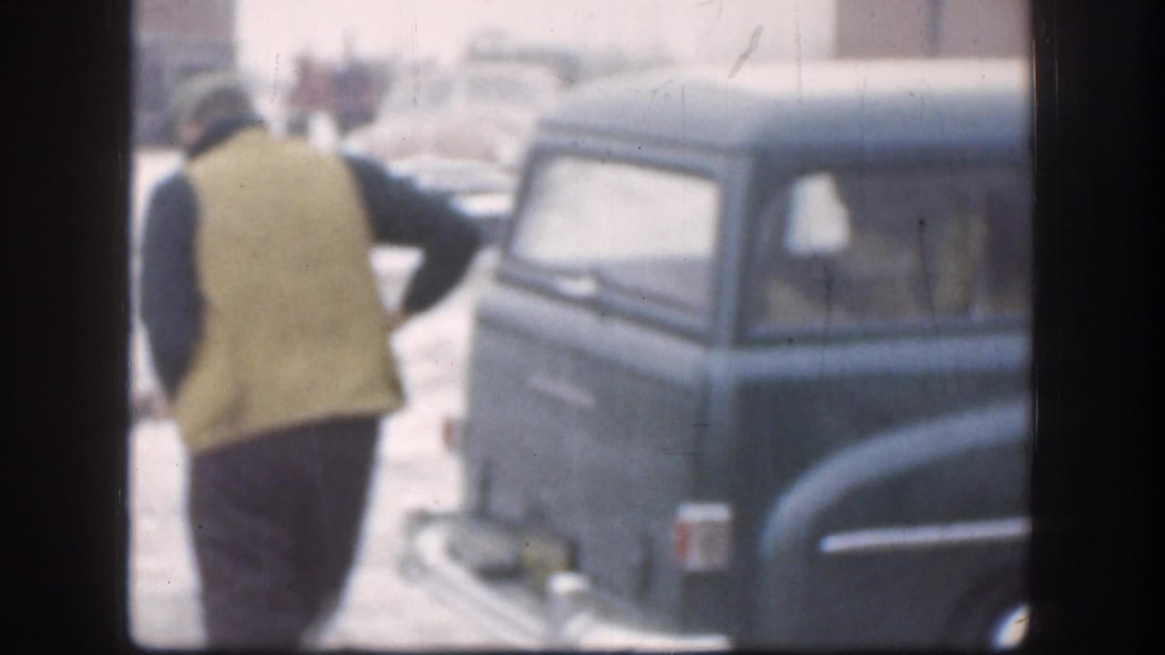 Kansas 1958 home-movie frame showing a person walking beside a rounded station wagon parked near the porch area.