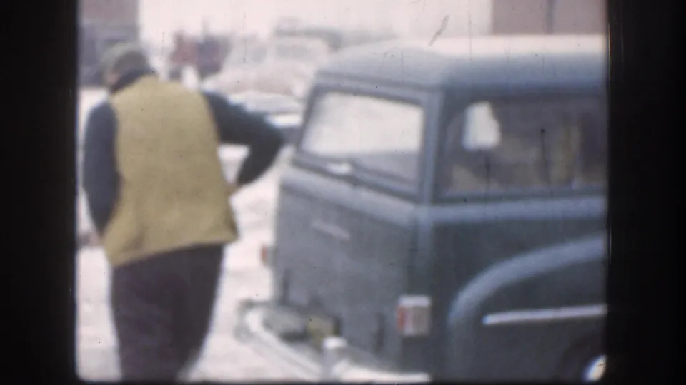 Kansas 1958 home-movie frame showing a person walking beside a rounded station wagon parked near the porch area.